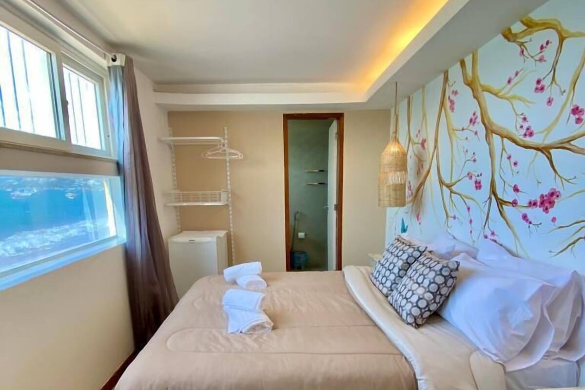A bedroom with a bed, a window with curtains, a wall-mounted shelf, and a door.