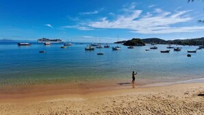 A beach with a person standing on the shore, boats in the water, and a cruise ship in the distance. - Sabatico Beach House (Búzios)
