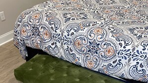 A bed with a patterned bedspread, a green upholstered bench, and a multicolored striped rug.