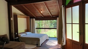 A bedroom with a wooden ceiling, a bed, a small table, and a view of greenery outside. - Loft's Jardim's - Campeche - Floripa (Florianópolis)