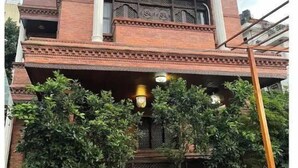 Ein mehrstöckiges Gebäude im traditionellen Baustil, mit einem Balkon im zweiten Stockwerk und einem dekorativen Eingangsbereich mit kleinen Gartenanlagen davor.
