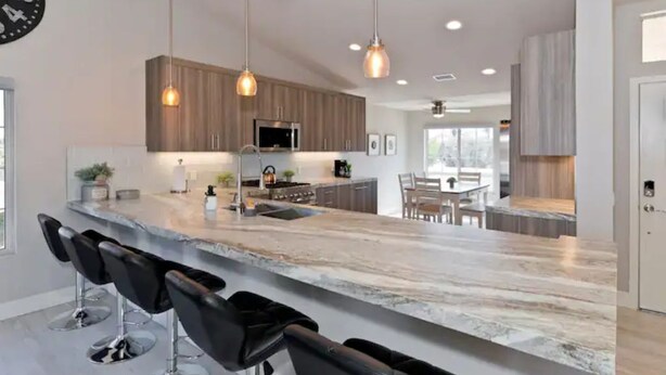 A modern kitchen with a marble countertop, bar stools, pendant lighting, and wooden cabinets. - Luxury Getaway - Casa Vaugeois (Palm Desert)