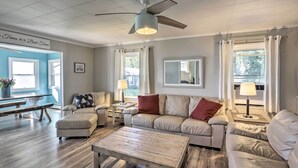 A spacious living room with a ceiling fan, a wooden coffee table, a beige sofa, and a dining table with chairs. - Lakefront Cottage w/ Fishing & Private Yard! (Port Clinton)