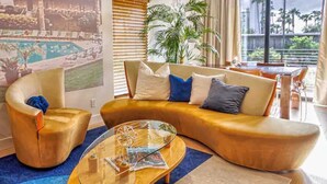 A modern living room with a curved sofa, a glass coffee table, and a view of a pool and palm trees through large windows.