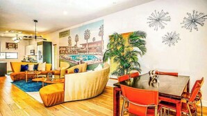 A modern living room with a sofa, coffee table, and dining table. There is a large painting on the wall and a potted plant.