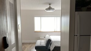 Una habitación con cama, ventilador de techo, ventana y refrigerador.