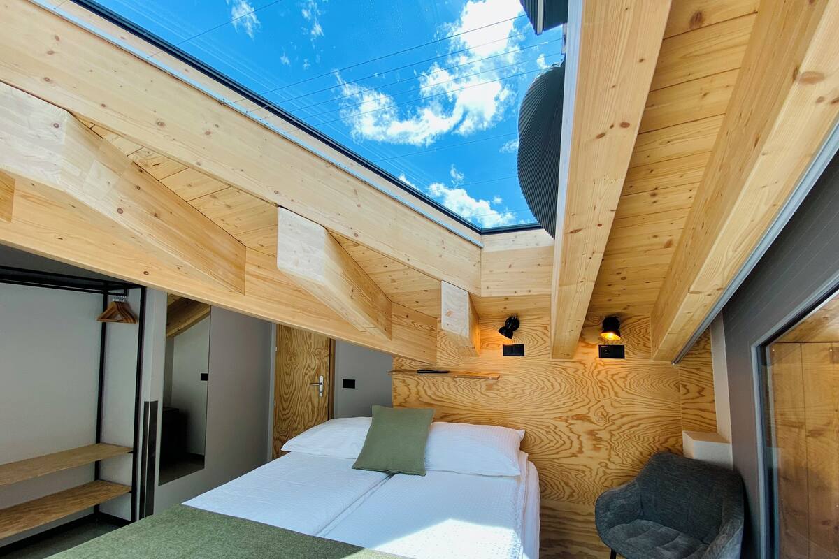 A compact bedroom with a skylight, a bed with pillows, a chair, and a small table.
