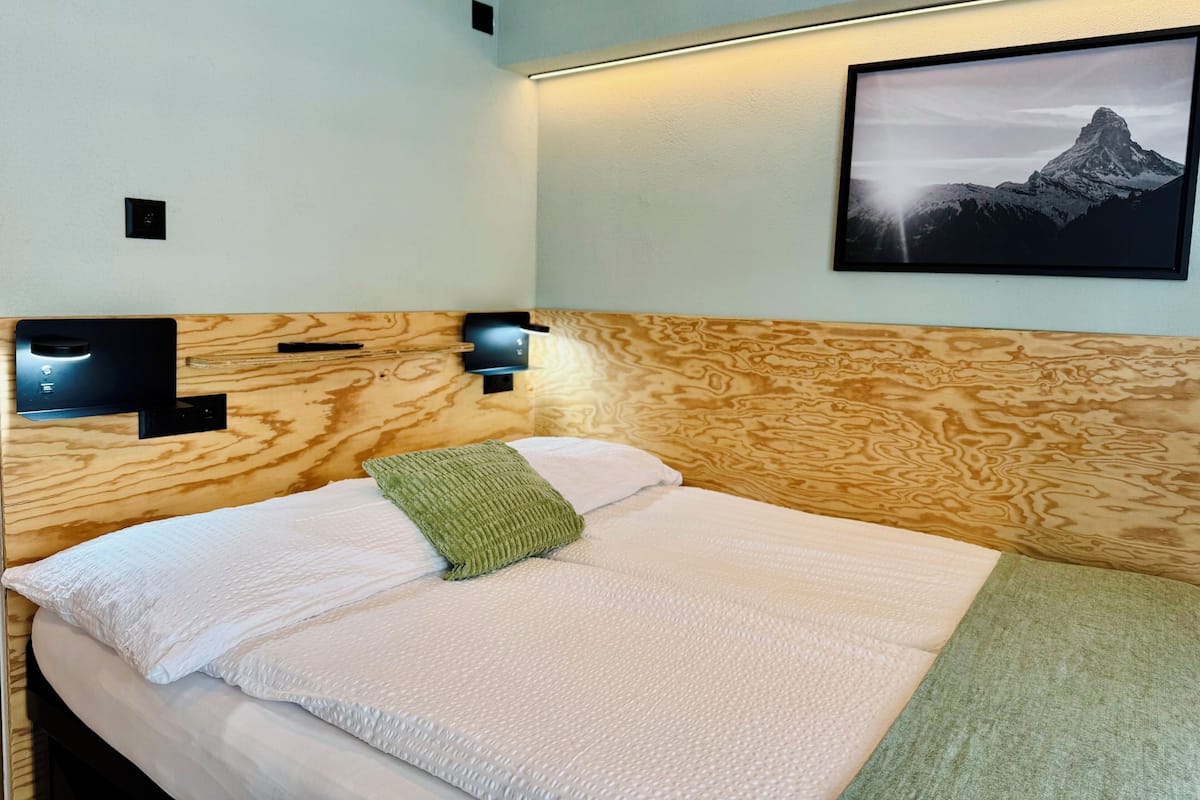 A neatly made bed with a green pillow and a white blanket, wooden headboard, two wall-mounted lights, and a framed picture of a mountain.