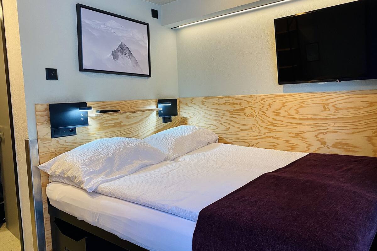 A neatly made bed with white linens and a dark throw blanket, a framed picture of a mountain on the wall, and a flat-screen TV mounted above the bed.
