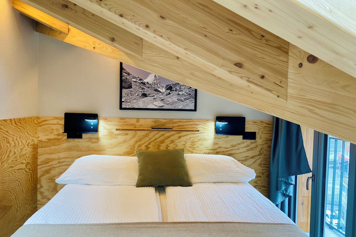 A wooden ceiling with exposed beams, a bed with white bedding and a green pillow, two wall-mounted speakers, and a framed picture on the wall.