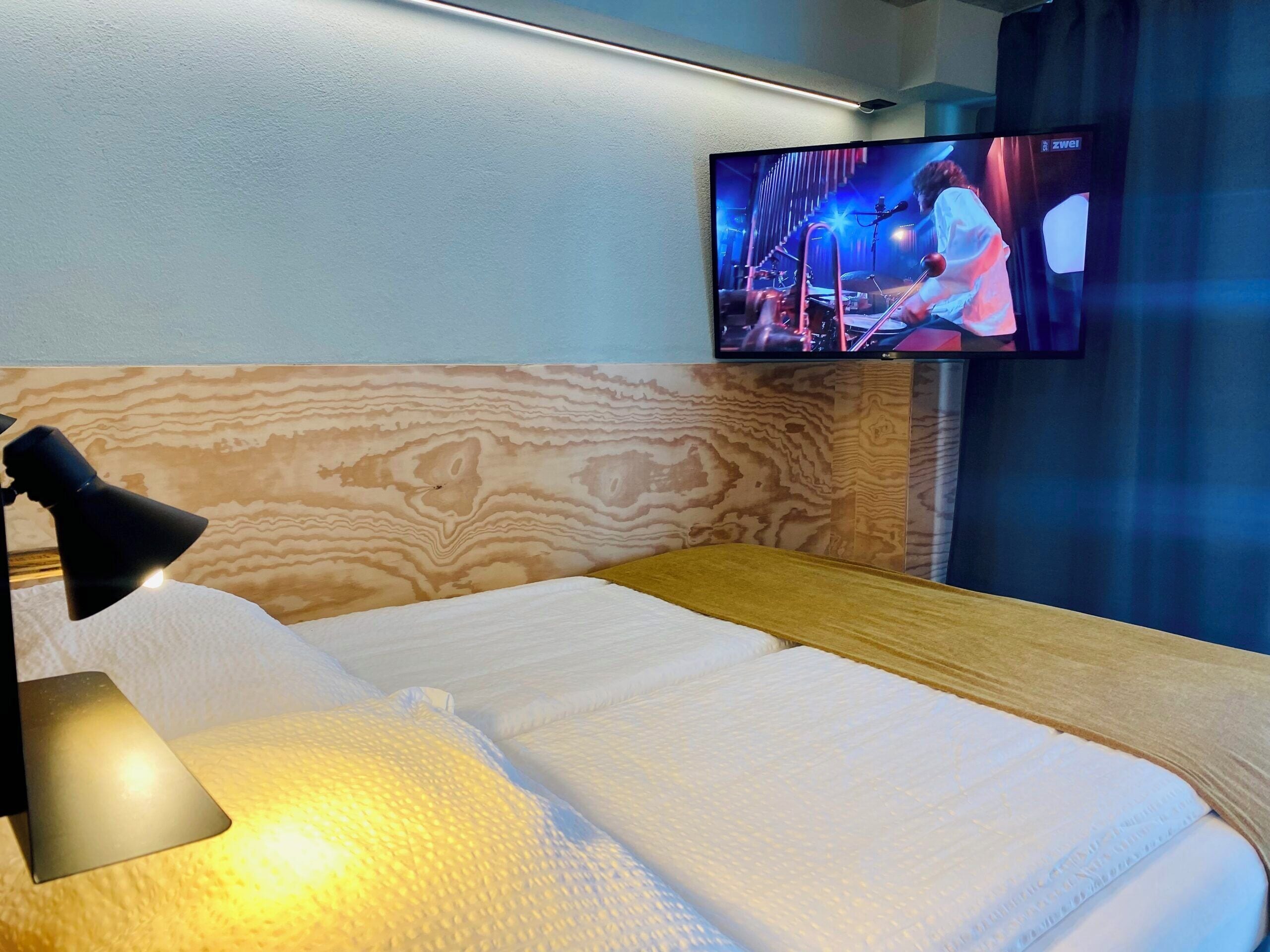 Habitación de hotel con una cama, una lámpara, un televisor montado en la pared y un cabecero de madera.