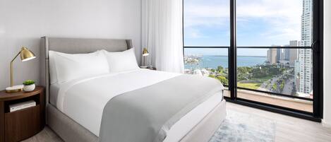 A modern hotel room with a large bed, a nightstand, a lamp, and a view of the city and water.