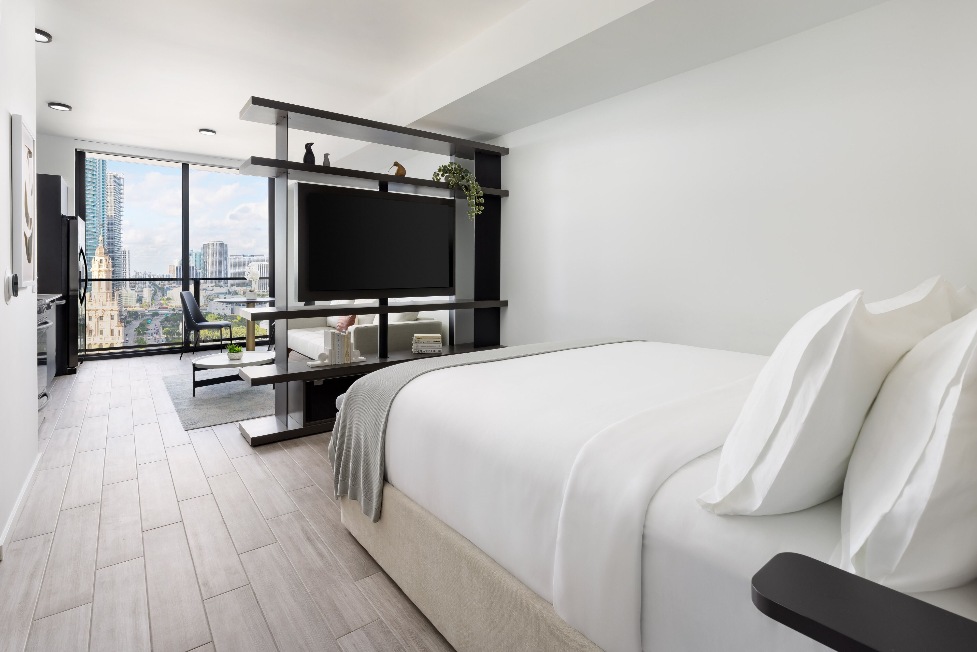 Una camera d'albergo moderna con un letto grande, una TV montata su una cornice nera, un balcone con vista sulla città e un'area pranzo con tavolo e sedie.