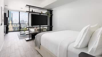 Una camera d'albergo moderna con un letto grande, una TV montata su una cornice nera, un balcone con vista sulla città e un'area pranzo con tavolo e sedie.