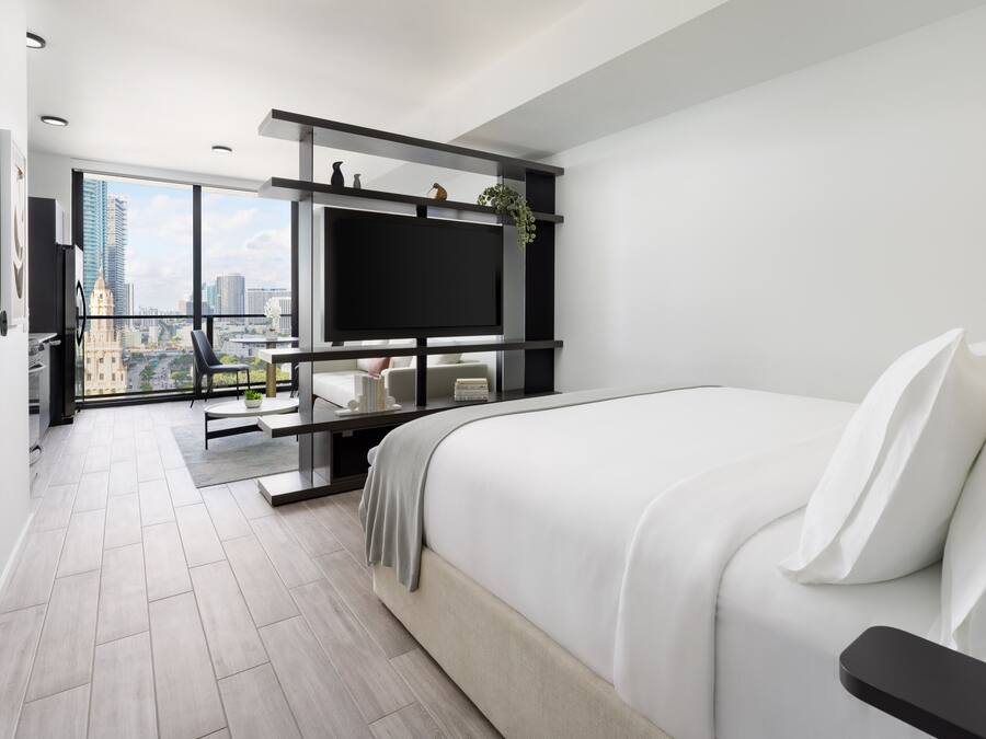 A modern hotel room with a large bed, a TV mounted on a black frame, a balcony with a city view, and a dining area with a table and chairs.