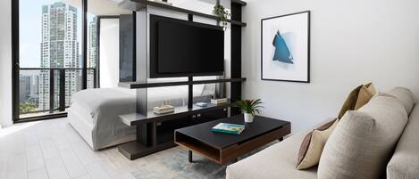 A modern living room with a large flat-screen TV mounted on a black shelf, a beige sofa, a coffee table with a book, and a city view through large windows.