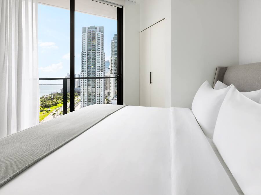 A modern hotel room with a large bed, a view of high-rise buildings, and a balcony.