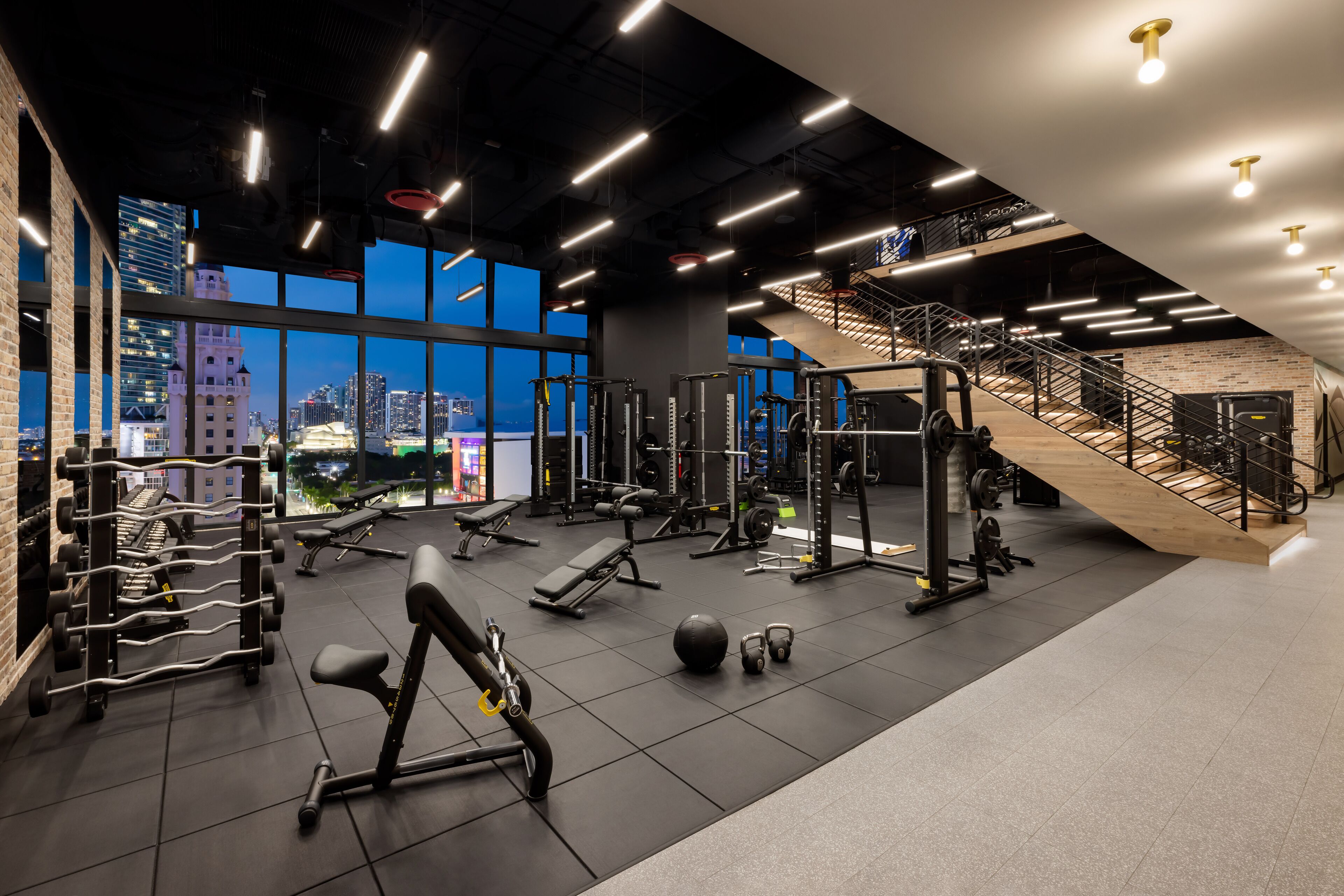 A modern gym with various exercise equipment, large windows offering city views, and a staircase leading to an upper level.