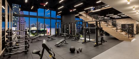A modern gym with various exercise equipment, large windows offering city views, and a staircase leading to an upper level.