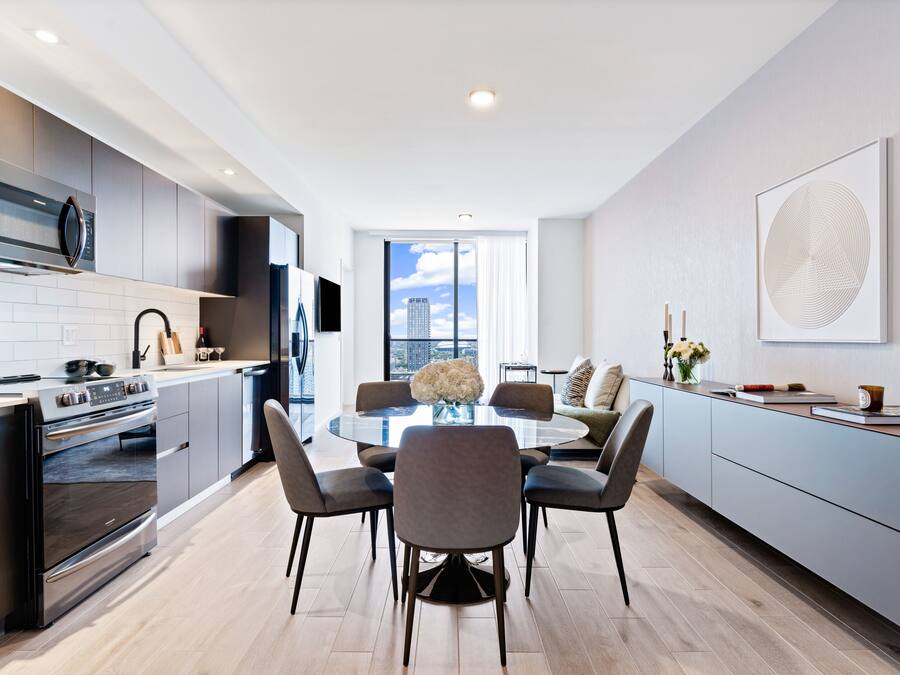 A modern kitchen with a dining area, featuring a central island, built-in appliances, and a large window offering a city view.