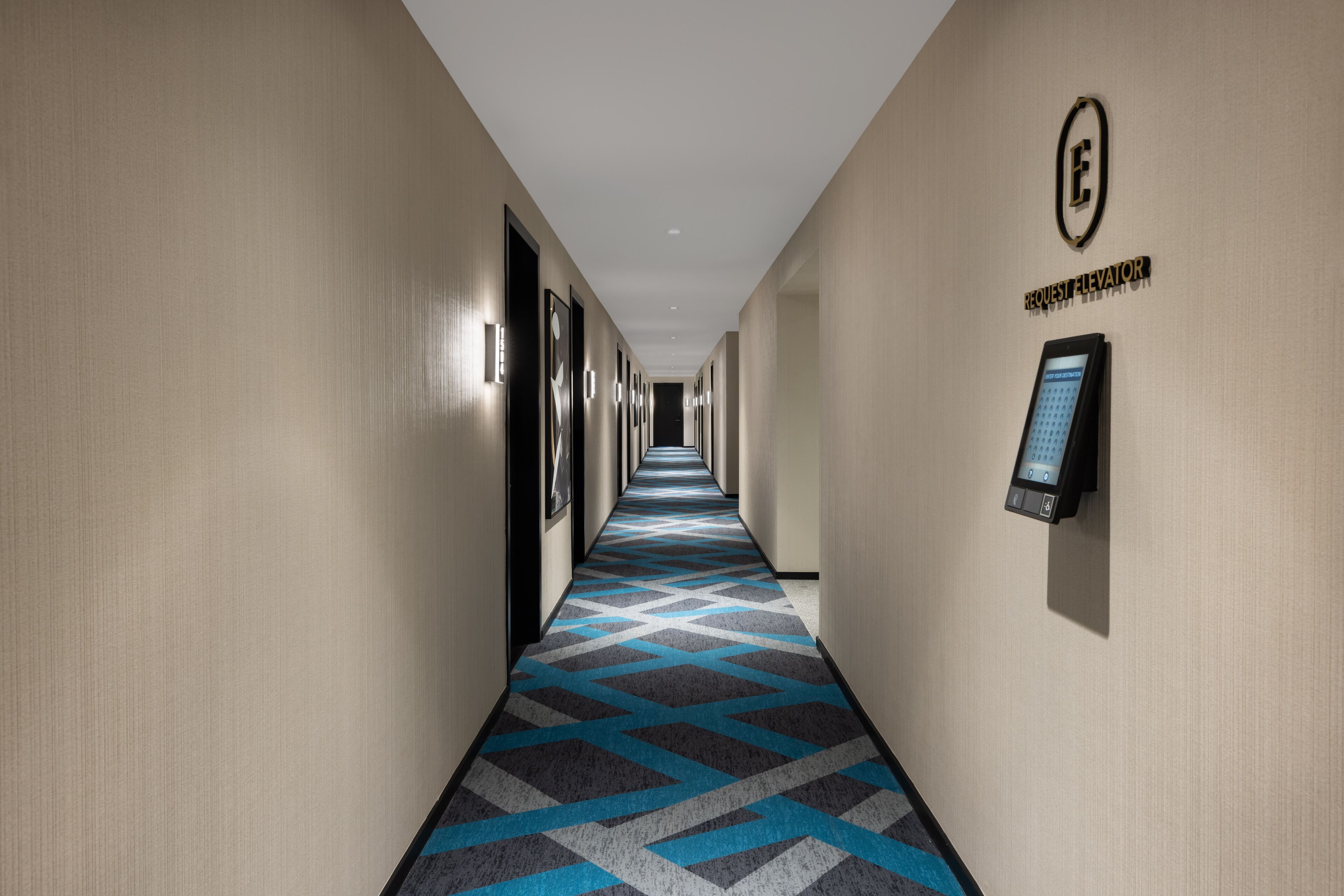 A hotel hallway with a patterned carpet, a request elevator sign, and a digital elevator control panel.