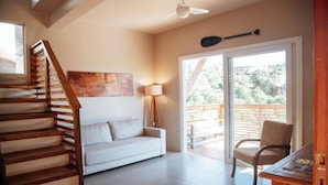 Een ruime woonkamer met een houten trap, een witte bank, een vloerlamp en een glazen schuifdeur die leidt naar een terras.
