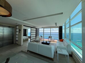Une chambre d’hôtel moderne dotée d’une grande fenêtre donnant sur la mer, d’un canapé blanc, d’une petite table et d’un espace nuit séparé avec un lit.