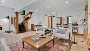 Amplia sala de estar con una escalera de madera, un comedor con sillas y una cocina con ventana.