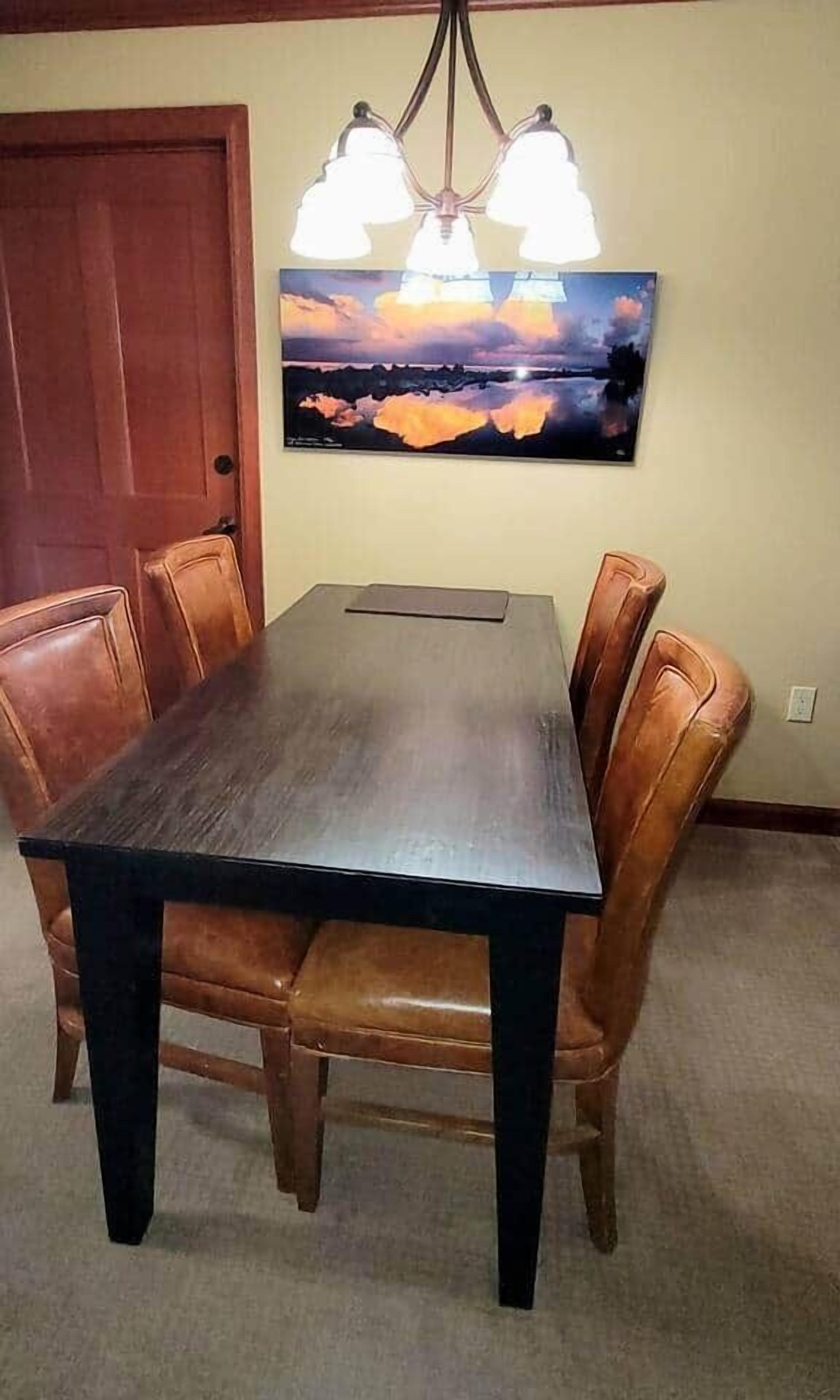 A dining table with six brown leather chairs, a wooden door, and a wall-mounted painting of a sunset over water.