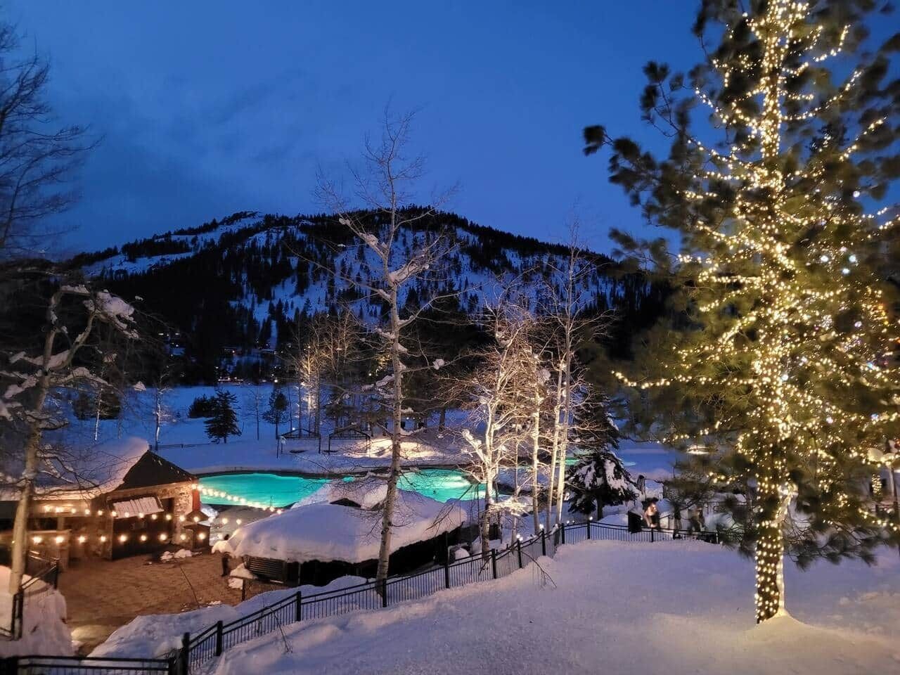 Un paysage enneigé avec une piscine, des arbres décorés de guirlandes lumineuses et une montagne en arrière-plan.