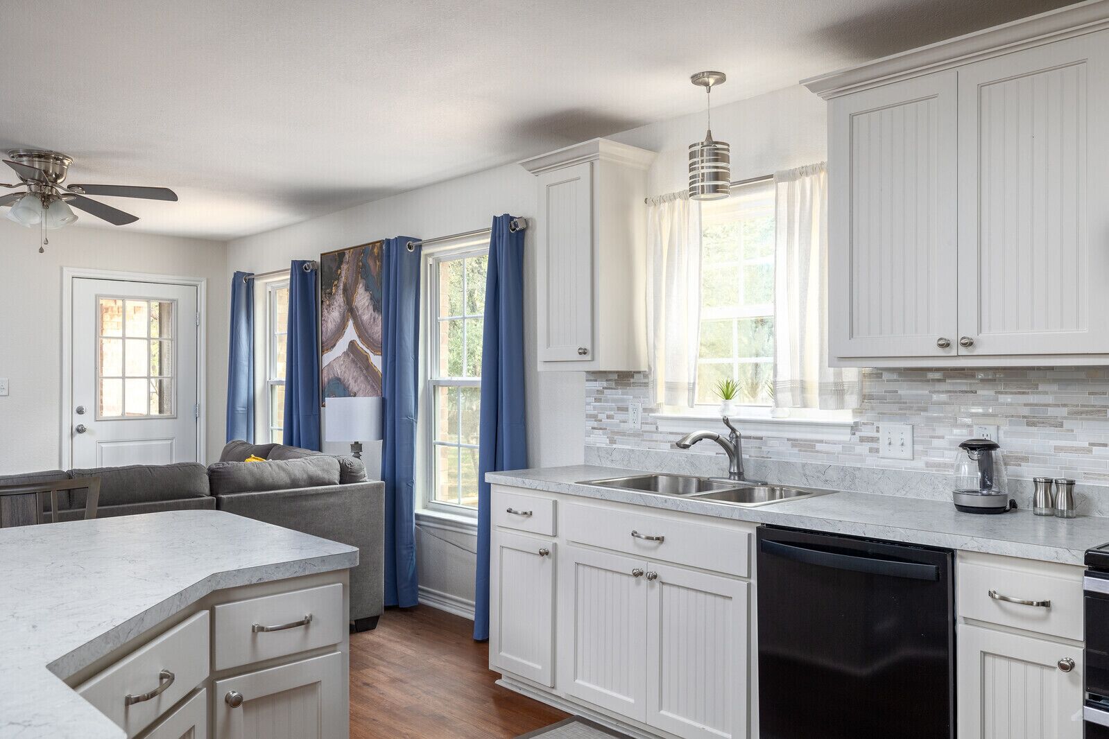 A modern kitchen with white cabinetry, a marble countertop, a built-in dishwasher, and a window with blue curtains.