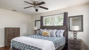 Un dormitorio con cama, ventilador de techo, ventana con cortinas, mesita de noche con lámpara y cómoda.