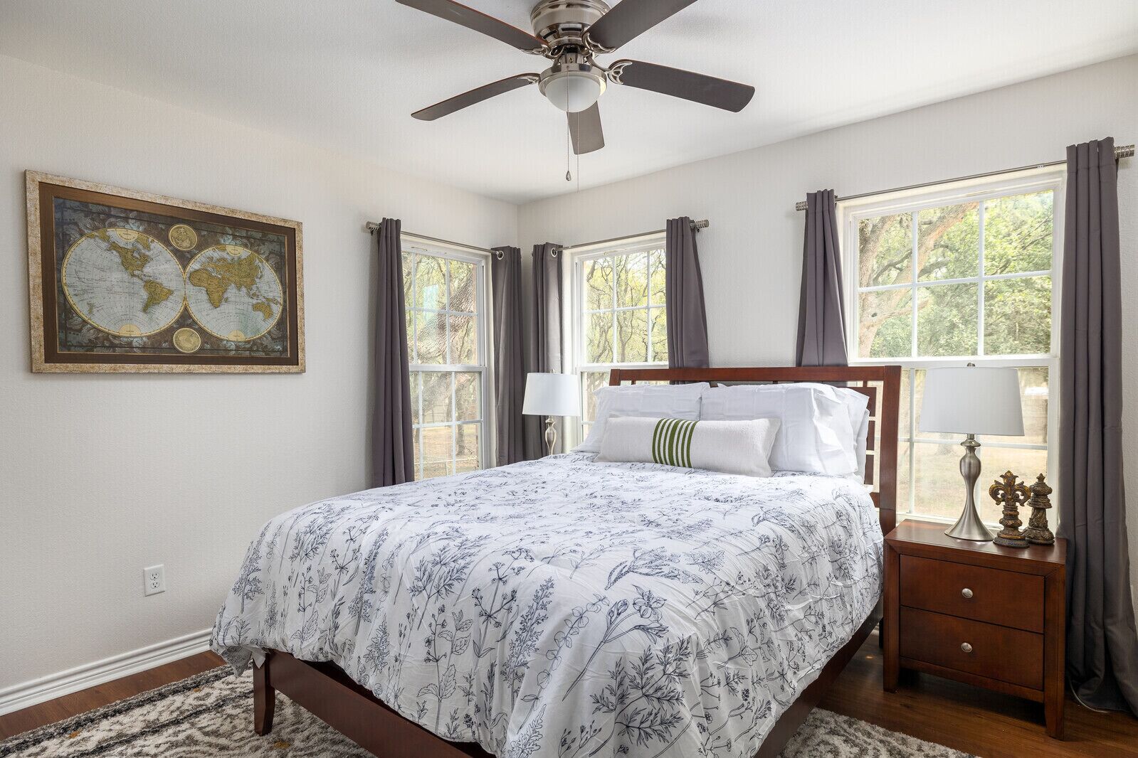 Une chambre à coucher avec un lit, un ventilateur de plafond, une table de chevet avec une lampe et une grande fenêtre avec des rideaux.