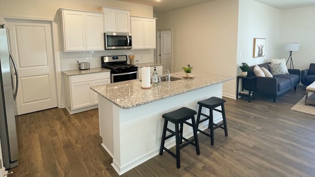 A modern kitchen with white cabinetry, a granite countertop, and stainless steel appliances. There is a small island with two stools and a living area with a dark sofa and a lamp. - Beautiful new home with lake view near hiking trails (Oak Ridge)
