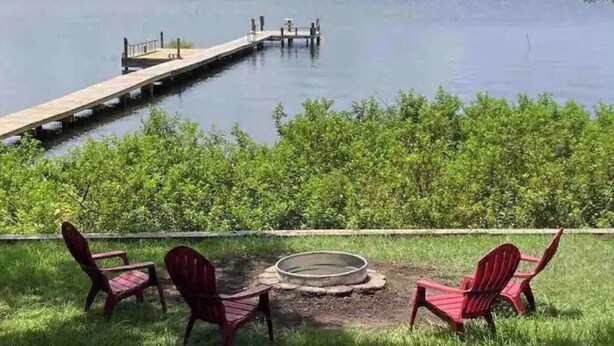 Vista lacustre con un muelle que se adentra en el agua, rodeado de vegetación exuberante y sillas Adirondack rojas orientadas a una fogata.