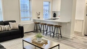 Un salon moderne avec un canapé noir, une table basse en bois et un coin cuisine équipé de tabourets de bar.