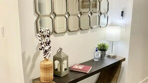 Un couloir moderne avec un mur décoré d’un miroir aux motifs géométriques, une console en bois et une lampe.