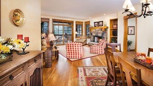Un salon chaleureux avec une cheminée, des meubles en bois et une vue sur l’extérieur grâce à de grandes fenêtres.