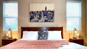 Une chambre d’hôtel avec un grand lit, deux tables de chevet avec des lampes, un tableau représentant un paysage urbain accroché au mur et une fenêtre avec des stores.