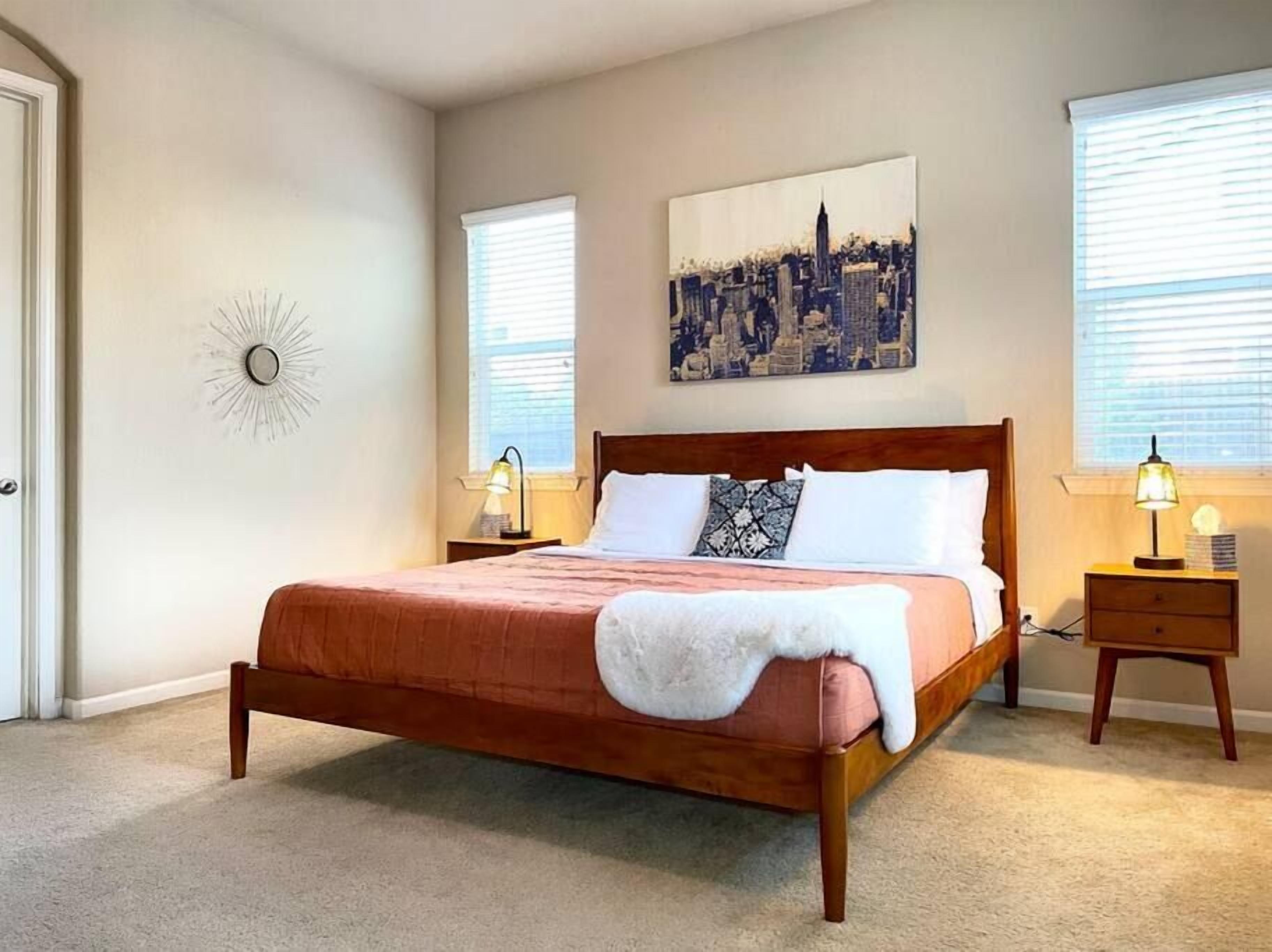 Une chambre avec un lit en bois, deux tables de chevet avec des lampes, un tableau représentant un paysage urbain accroché au mur et un élément décoratif mural en forme de soleil rayonnant.