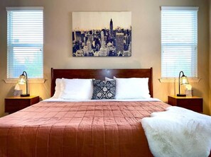 A bedroom with a cityscape painting, a wooden headboard, a pink quilted bedspread, two bedside lamps, and a white throw blanket.