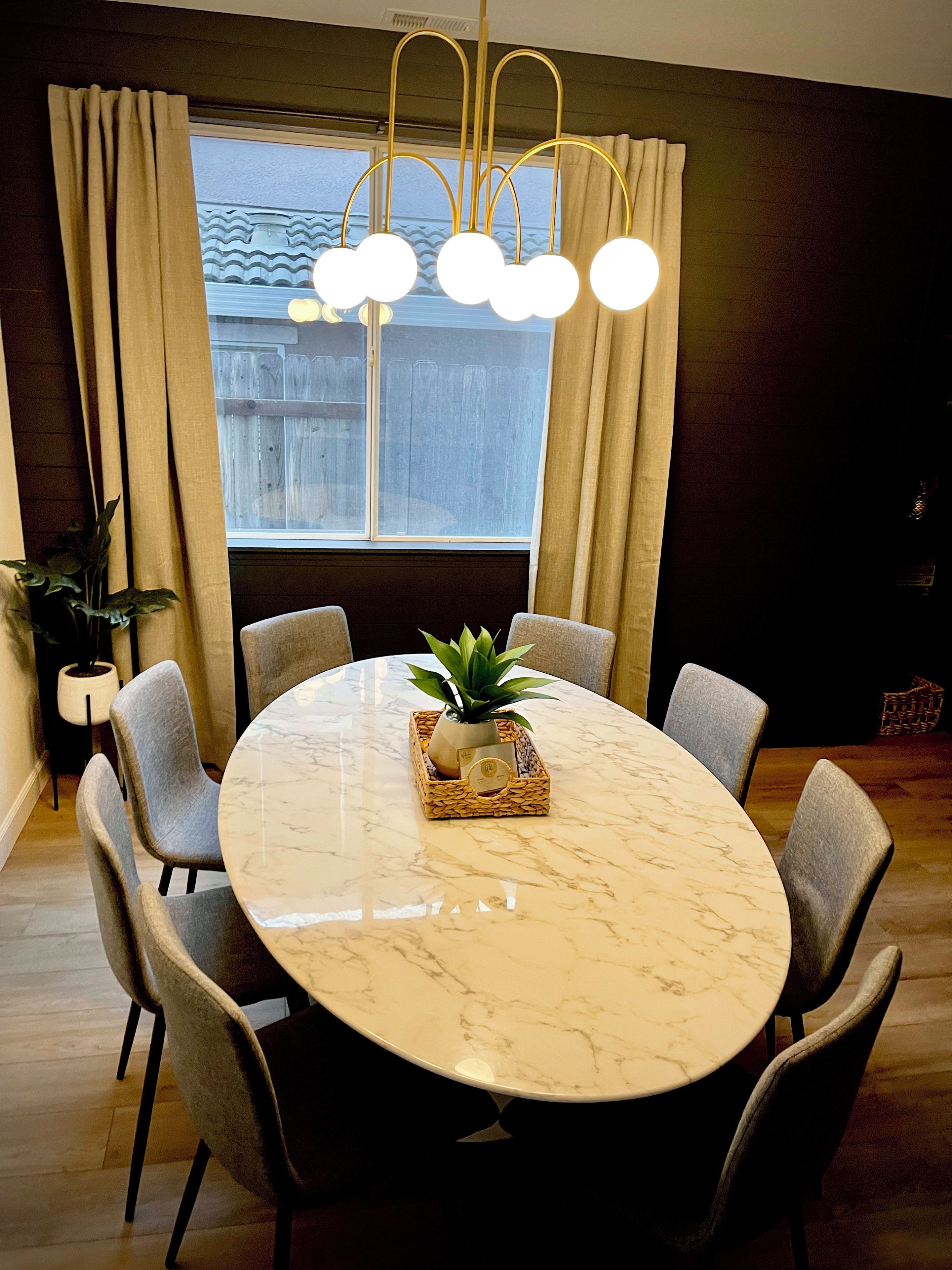Uma sala de jantar com uma mesa redonda de mármore, seis cadeiras cinzas, uma planta central e um lustre moderno.