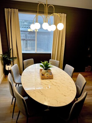 Een eetgedeelte met een ronde marmeren tafel, zes grijze stoelen, een plant in pot en een moderne kroonluchter.