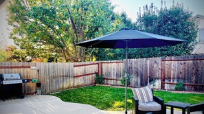 Un patio trasero con cerco de madera, una parrilla, una sombrilla y mobiliario exterior.