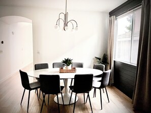 A dining area with a round table, six chairs, a hanging light fixture, and a window with curtains.