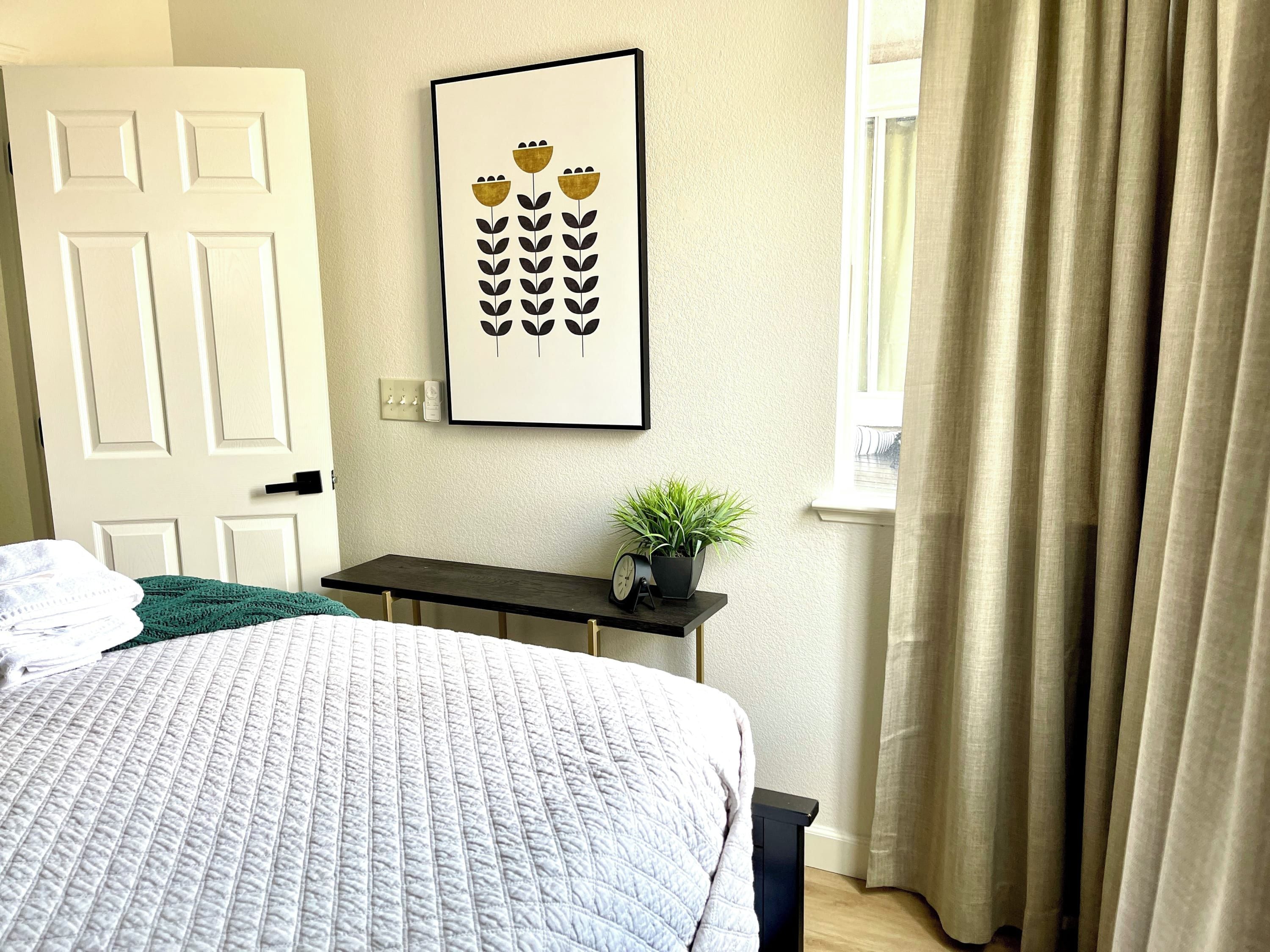 Un dormitorio con cama, una mesita de noche con una planta, un cuadro en la pared y una ventana con cortinas.