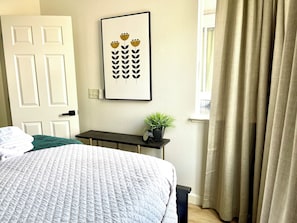 Un dormitorio con cama, una mesita de noche con una planta, un cuadro en la pared y una ventana con cortinas.