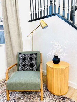Una habitación con una butaca tapizada de verde, una mesita de madera con un florero y una lámpara de pie.
