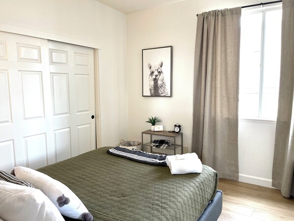 Une chambre à coucher avec un lit, des oreillers, une table de chevet avec une lampe, un tableau encadré et une fenêtre avec des rideaux.