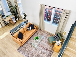 Un salon avec un canapé, une table basse et un fauteuil. Il y a une fenêtre avec des rideaux, une plante et une vue sur un bâtiment à l’extérieur.
